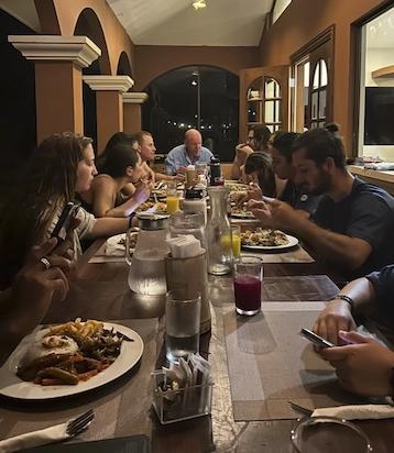 Students and researchers sit at a long table with dinner plates full of food in front of them. Some people are eating, others are talking to each other.