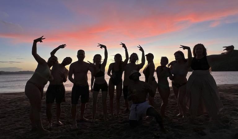 Studetns and researchs stand shoulder to shoulder waving and throwing up peace signs at the camera. They are all covered in shadows while a beautiful sunset with pink clouds sits behind them.
