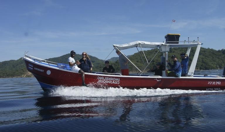 Students and researchers smile and wave at the camera. They're on a red boat with white trim that is sailing on water.